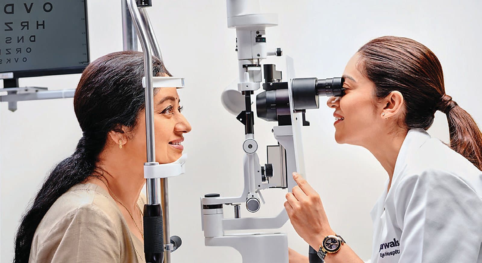 A Dr Agarwals ophthalmologist examining a patient at a slit lamp, a letter chart on the wall behind them